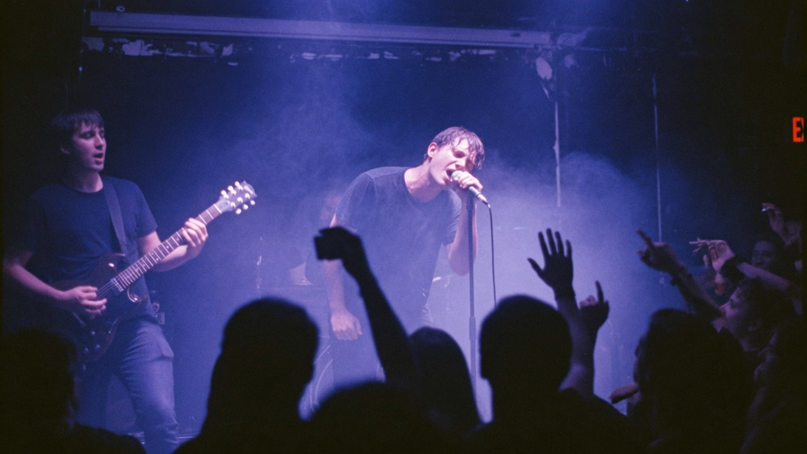Audience view of a vocalist and guitarist on a hazy club stage under blue and purple lights, with hands raised in the crowd