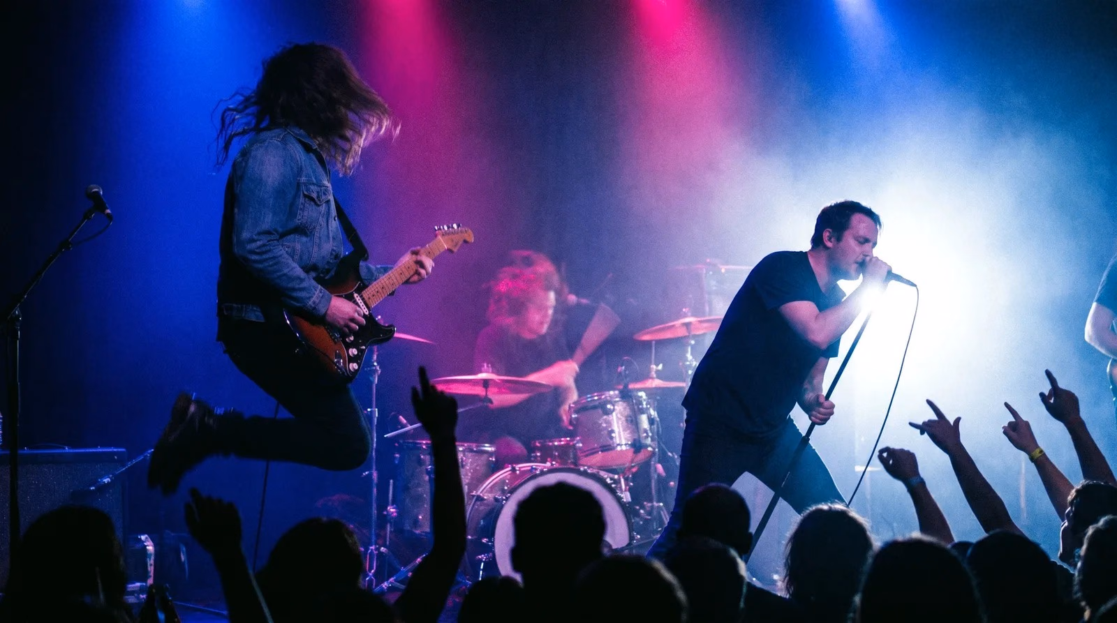 Live band on stage with a guitarist mid-jump, a vocalist at the mic, and crowd hands raised under blue and magenta lights